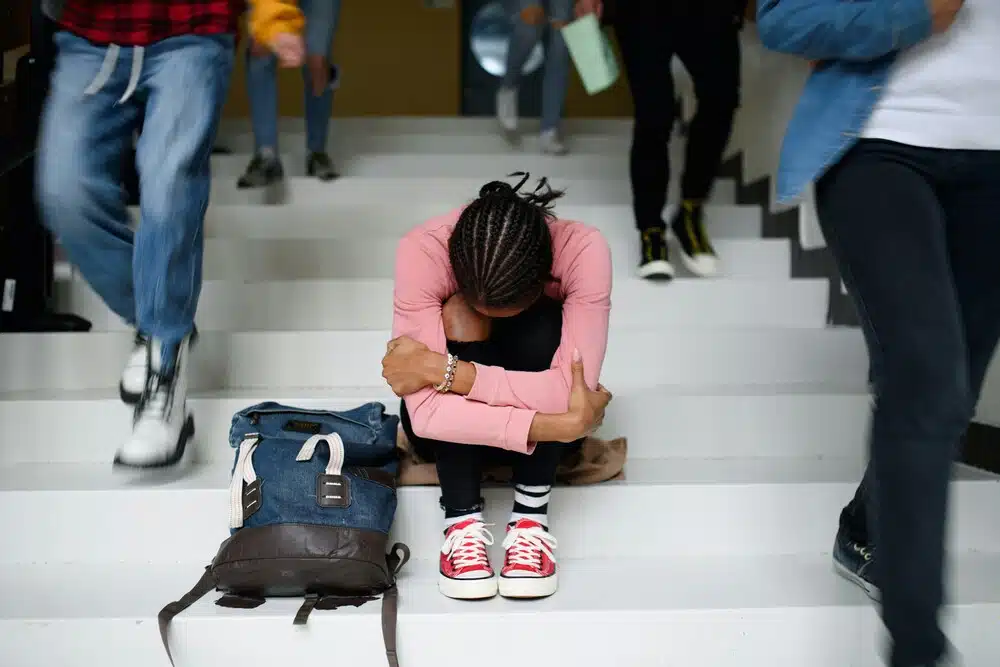 Teenager sitting alone on school stairs with head down while other students walk past, illustrating social withdrawal and warning signs of depression in teens