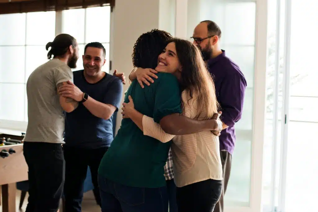 A warm gathering of friends in a sunlit room, focusing on a woman smiling brightly as she hugs a friend, while others greet each other in the background—symbolizing the importance of having a robust social circle outside of a primary partnership.