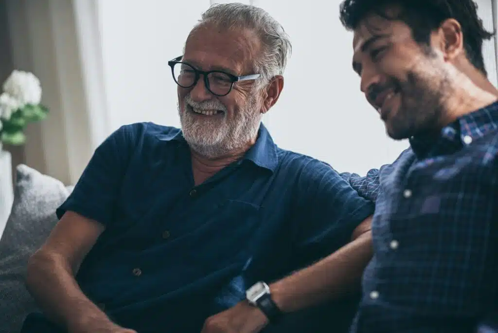 A close-up of a relaxed, grey-bearded older man wearing glasses smiling warmly next to a smiling younger man on a couch, capturing the sense of relief and connection that comes from unburdening life's pressures.