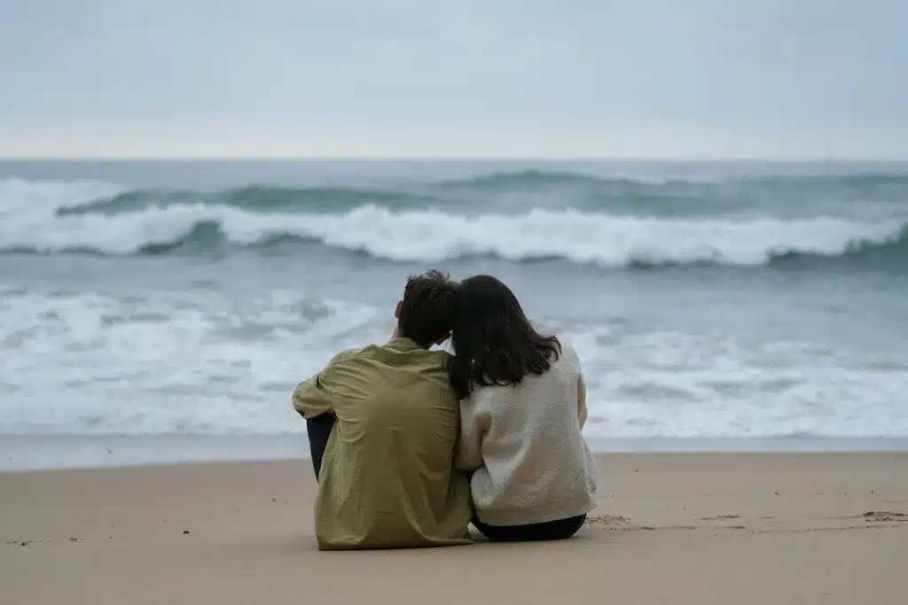A rear view of a couple sitting close together on a sandy beach, looking out at crashing ocean waves under an overcast sky.