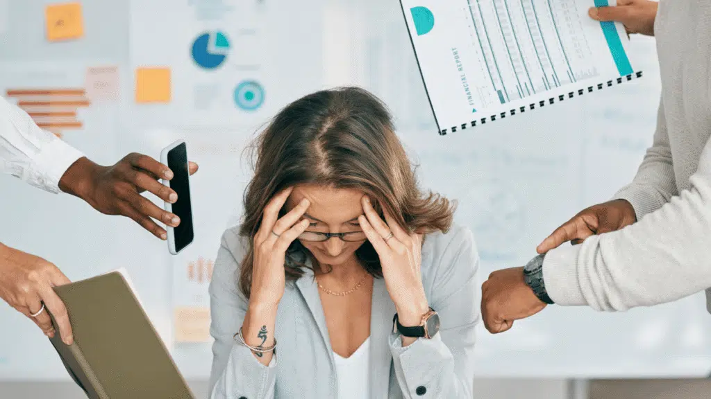 Woman holding her head at a desk as colleagues hand her a phone, files, and reports, overwhelmed by constant workplace demands.
