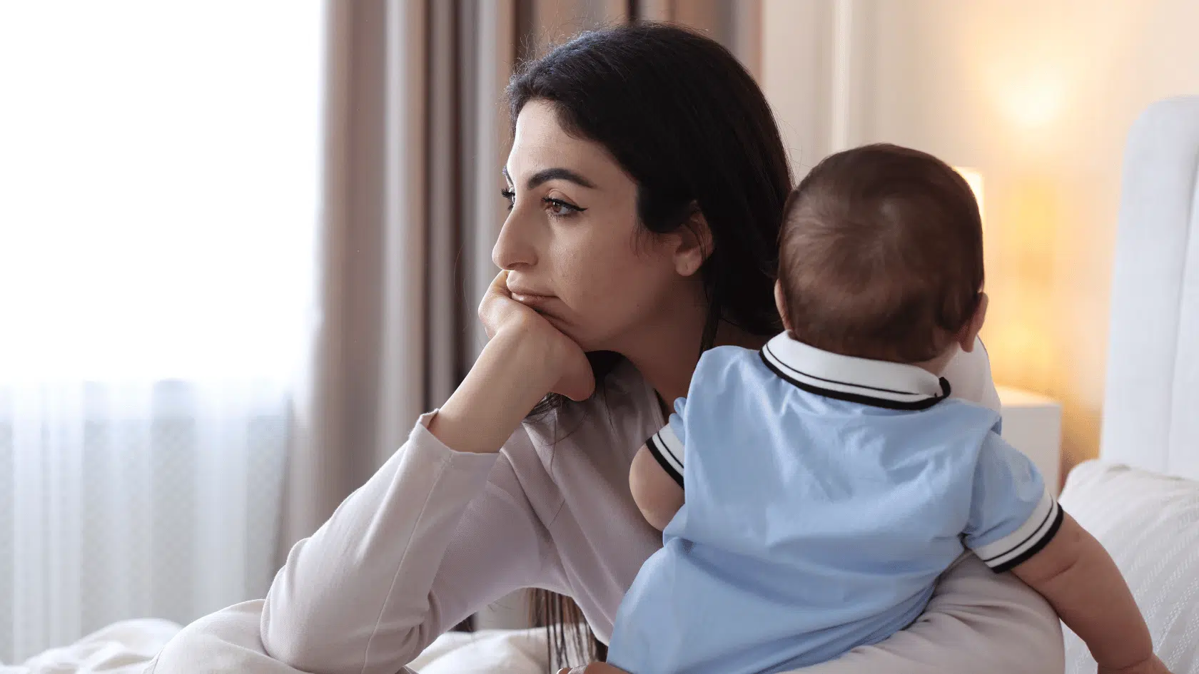 Mother holding her baby and gazing out a window with a heavy expression, reflecting the emotional weight of postpartum depression.