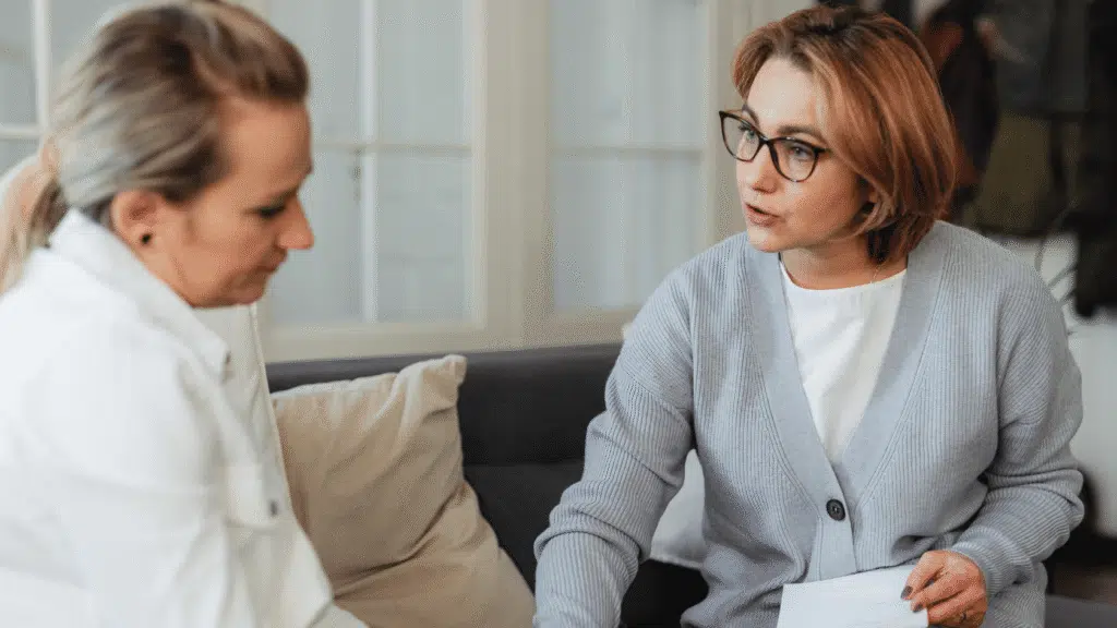 Counselor holding notes and explaining her therapeutic approach to a client seated beside her during an intake conversation.