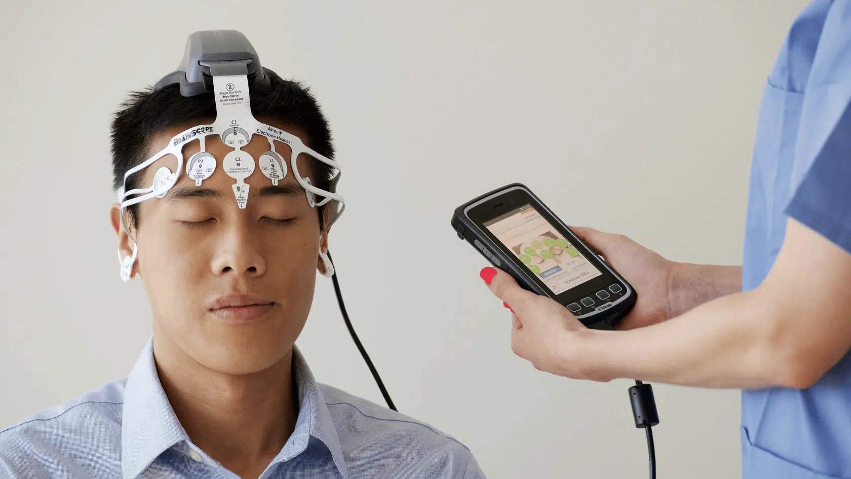 Man with his eyes closed wearing an EEG electrode headset while a clinician monitors brain activity readings on a handheld device.