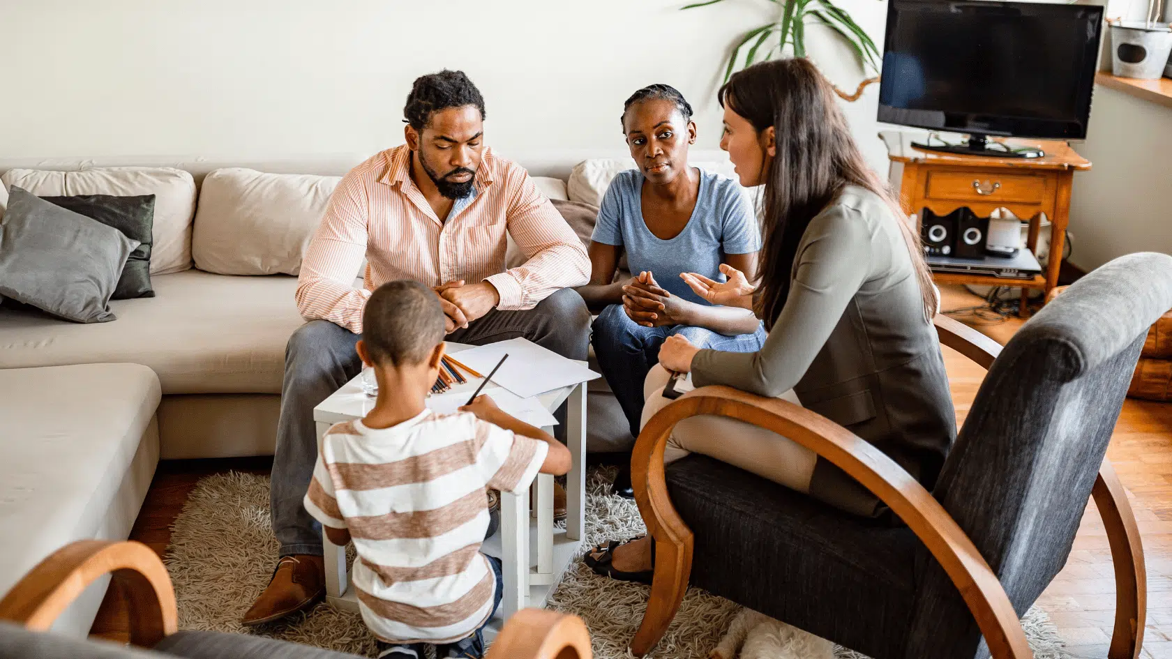Couple sitting with a counselor in a living room while their young child draws nearby, talking through postpartum struggles together.