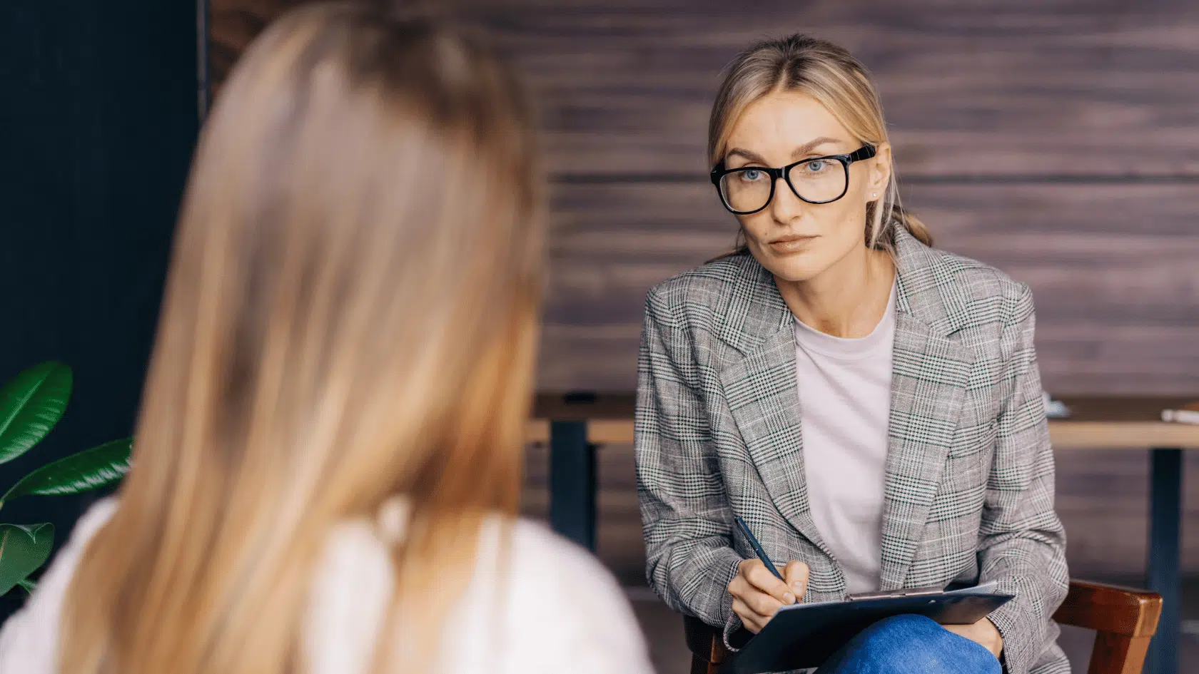 Therapist in glasses holding a notepad and listening attentively as a client speaks during a counseling session in a warm office.
