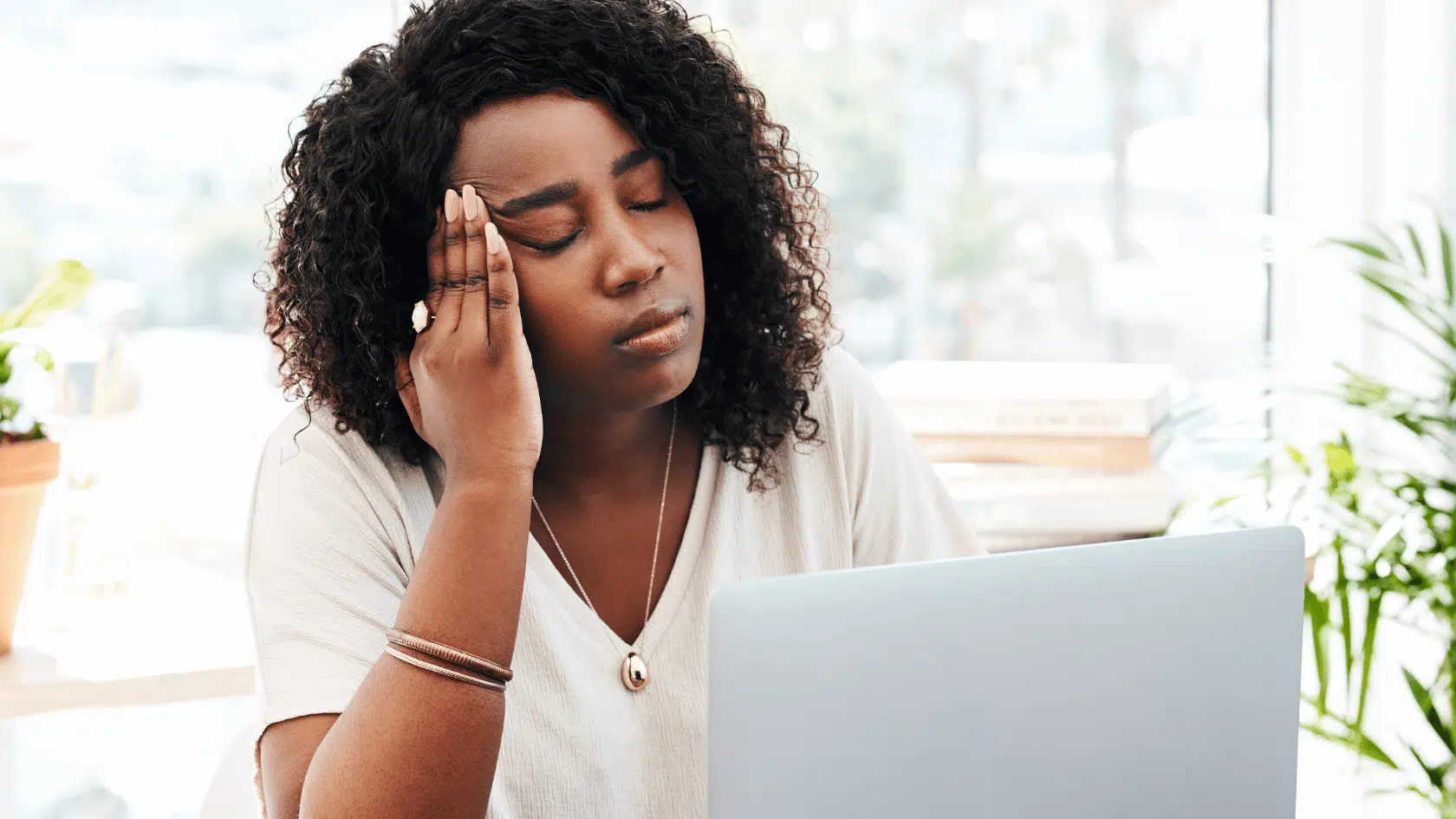 Woman sitting at her laptop with her eyes closed and hand to her head, showing the mental exhaustion that often signals burnout.